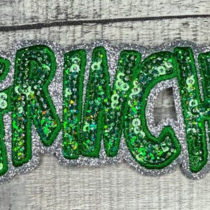 May include: A sequined patch with the word "GRUNCHY" in green letters outlined in silver glitter. The letters are filled with green sequins, creating a sparkling effect. The patch is on a gray surface.