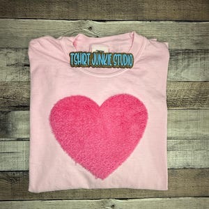 May include: A light pink t-shirt with a large, textured, bright pink heart in the center. The text "TSHIRT JUNKIE STUDIO" is printed on a label near the collar. The shirt is folded neatly on a wooden surface.