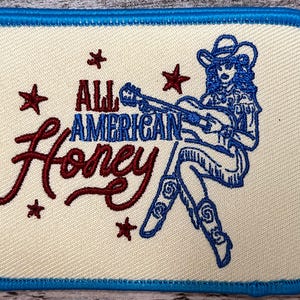 May include: Embroidered patch with a blue border and a white background. The patch features a woman playing a guitar with the text "All American Honey" in red.