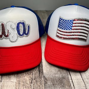 May include: Two white trucker hats with red and blue accents. One hat features a sparkly red, white, and blue American flag. The other hat features the word "USA" in sparkly red, white, and blue letters.