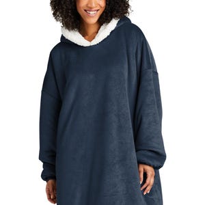 May include: A navy blue oversized hooded blanket with a white sherpa-lined hood. The blanket features long sleeves and a relaxed fit. The model is wearing the blanket. This cozy wearable blanket is designed for comfort and warmth.