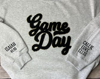 Può includere: Una felpa grigia con le parole "Game Day" in nero con accenti di glitter dorati. La manica sinistra ha il testo "Isaiah 41:10" e la manica destra ha il testo "Callie Luke Ellis".