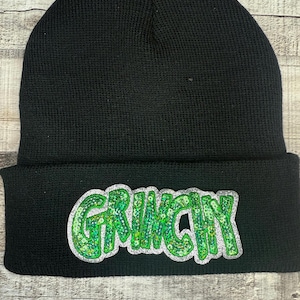 Puede incluir: Gorro de punto negro con la palabra "GRINCHY" en lentejuelas verdes y purpurina plateada. El gorro está doblado en el borde y se encuentra sobre una superficie de madera.