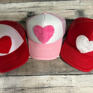May include: Three trucker hats with heart designs. One hat is pink with a pink heart, and the other two are red with red and white hearts. The hats have a mesh back and a curved brim.
