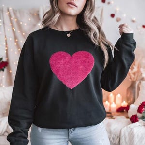 May include: Black sweatshirt featuring a large, textured, pink heart design on the front. The long-sleeved top is made of a soft material. The background includes candles and decorative elements, suggesting a Valentine's Day theme.