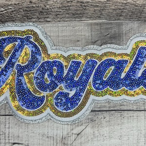 May include: A sequined patch with the word "Royals" in blue script. The letters are filled with blue sequins and outlined with gold and silver sequins. The patch is on a wooden background.