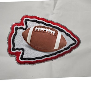 May include: A brown football with white stripes rests on a white background within a red, black, and silver arrowhead design. The football has white laces and is centered in the image.