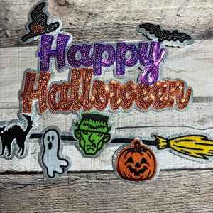 May include: A collection of Halloween-themed patches. The patches include a witch's hat, a bat, a black cat, a ghost, Frankenstein's monster, a jack-o'-lantern, and a broom. The words "Happy Halloween" are written in sequined letters.