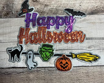 Puede incluir: Un conjunto de parches con temática de Halloween. Los parches incluyen un sombrero de bruja, un murciélago, un gato negro, un fantasma, el monstruo de Frankenstein, una calabaza y una escoba. Las palabras "Happy Halloween" están escritas con letras de lentejuelas.