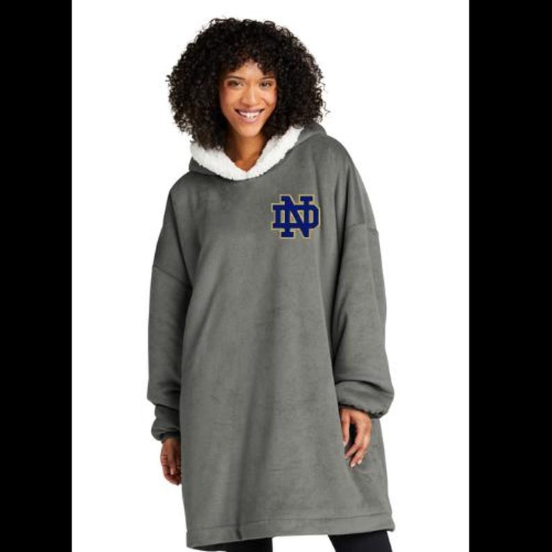 May include: A gray oversized hooded sweatshirt with a white sherpa-lined hood. The sweatshirt features a navy blue and gold embroidered logo on the chest. The model is wearing the sweatshirt.