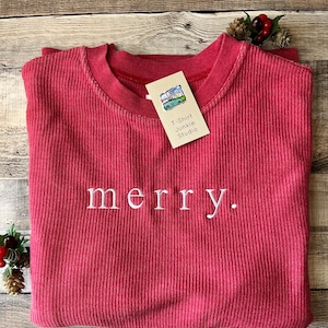 以下が含まれることがあります： 白い文字で「merry」と刺繍された赤いリブ編みのクルーネックセーター。 「T-Shirt Junkie Studio」のタグが付いています。 セーターは木製の表面に折りたたまれており、装飾的な松ぼっくりとベリーが付いています。