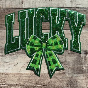 Op de afbeelding: Groene en witte glinsterende vilt applique met het woord "LUCKY" in een witte omtrek en een groen-witte geruite strik.