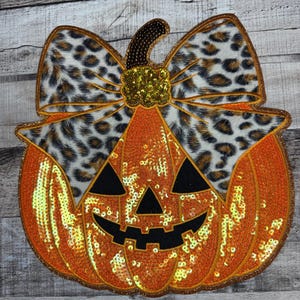 Peut inclure: Une citrouille orange à paillettes avec un visage de Jack-o'-lantern noir et un nœud léopard. La citrouille a un contour doré pailleté et une tige marron. Une décoration festive pour Halloween.