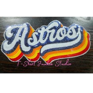 May include: A sequined Astros logo patch in white, blue, orange, and red. The word "Astros" is in a stylized font, outlined in white, with a blue sequin fill. The bottom of the patch has a rainbow design.