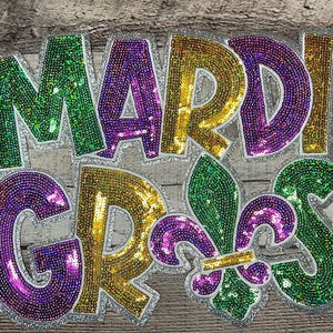 May include: A decorative sign with the words "MARDI GRAS" in large, sequined letters. The letters are in green, gold, and purple, with a silver glitter border. A fleur-de-lis is also included in the design.