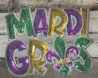 May include: A decorative sign with the words "MARDI GRAS" in large, sequined letters. The letters are in green, gold, and purple, with a silver glitter border. A fleur-de-lis is also included in the design.