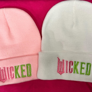 以下が含まれることがあります： 「WICKED」という単語が前面に刺繍されたニットのビーニーが2つ。1つはピンク、もう1つは白。ピンクのビーニーには「WICKED」という単語がピンクとグリーンで刺繍され、白いビーニーにはピンクとグリーンで刺繍されている。