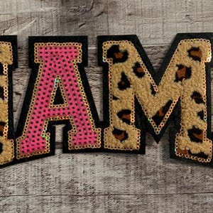 Peut inclure: Le mot "MAMA" est écrit en grandes lettres colorées. Les lettres sont en tissu avec un imprimé léopard et des paillettes roses.