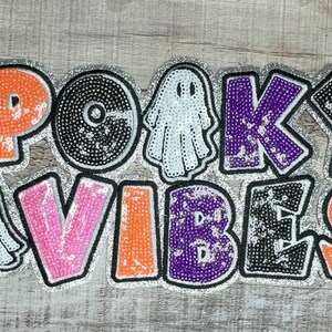 Op de afbeelding: Een met pailletten versierde Halloween-decoratie met de woorden "SPOOKY VIBES" in roze, oranje, zwart en paars. De letters zijn omlijnd met zwarte en zilveren glitter. Twee spookfiguren zijn ook in het ontwerp opgenomen.