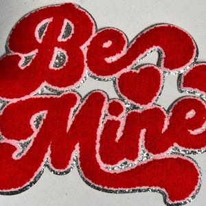 May include: A red felt applique with the words "Be Mine" in a script font. The letters are outlined in pink and silver glitter. A small pink heart is between the words. The applique is likely for Valentine's Day.