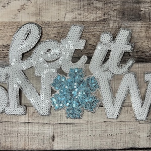 Puede incluir: ¡Que nieve! Un letrero blanco brillante con las palabras "Let it Snow" y un copo de nieve azul brillante en el centro.