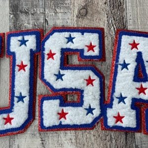 May include: White, blue, and red USA letter patches. Each letter is white with red and blue star accents. The letters are outlined in blue and red glitter. The patches are likely for apparel or crafts.