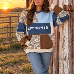Puede incluir: Una sudadera de manga larga con un diseño de patchwork de paneles de tela en azul claro, azul oscuro, marrón y crema, mezclados con un estampado de cachemira multicolor. El centro del pecho muestra la palabra carhartt en blanco.