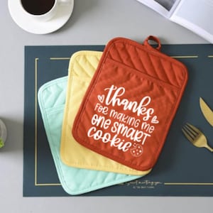 May include: Three quilted pot holders in red, yellow, and green. The red pot holder has the text "Thanks for making me one smart cookie" in white with a heart and a cookie graphic.
