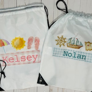May include: Two white drawstring bags with personalized names. The bag on the left has a sun, umbrella, and flip flops. The bag on the right has a ship, a wheel, and a map. Both bags have a checkered pattern.