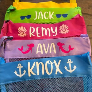 May include: A stack of colorful mesh beach bags with personalized names: Jack, Remy, Ava, and Knox. Each bag has a different color, including green, pink, purple, and blue. The names are in white, with decorative icons like sunglasses, seashells, mermaids, and anchors.