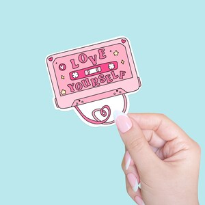 May include: Pink cassette tape sticker with the text "LOVE YOURSELF" in pink letters. The sticker has a white heart shape attached to it.
