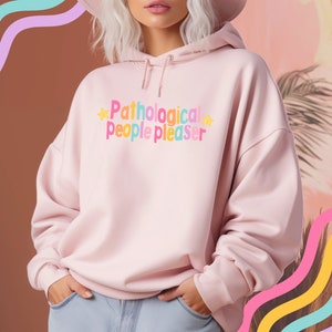K&ouml;nnte beinhalten: Ein rosa Hoodie mit dem Text "Pathological people pleaser" in Regenbogenfarben und gelben Sternen auf der Vorderseite.