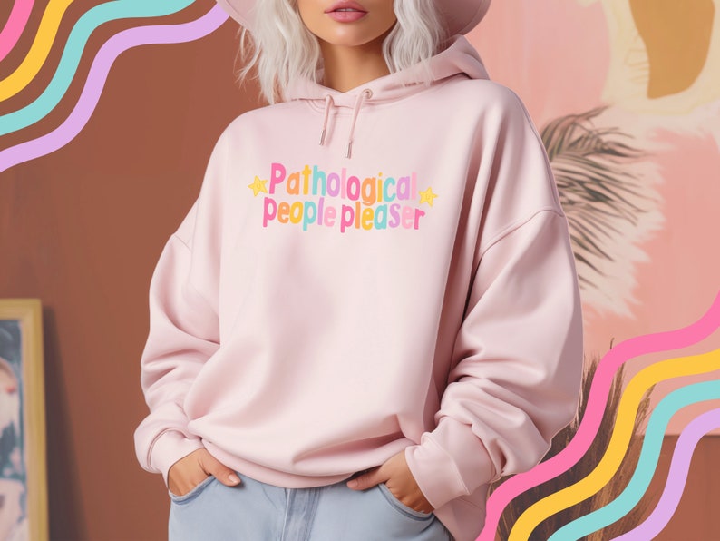 K&ouml;nnte beinhalten: Ein rosa Hoodie mit dem Text "Pathological people pleaser" in Regenbogenfarben und gelben Sternen auf der Vorderseite.