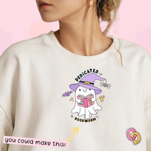 以下が含まれることがあります： 白いスウェットシャツに、紫色の魔女の帽子をかぶって本を読んでいる幽霊のグラフィックがプリントされています。幽霊の下には「Dedicated Bookworm」というテキストがあります。