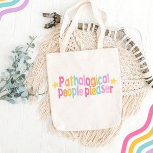 K&ouml;nnte beinhalten: Eine wei&szlig;e Canvas-Tragetasche mit dem Text "Pathological people pleaser" in bunten Buchstaben und gelben Sternen auf wei&szlig;em Hintergrund.