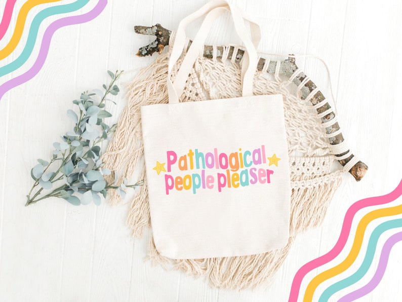 K&ouml;nnte beinhalten: Eine wei&szlig;e Canvas-Tragetasche mit dem Text "Pathological people pleaser" in bunten Buchstaben und gelben Sternen auf wei&szlig;em Hintergrund.