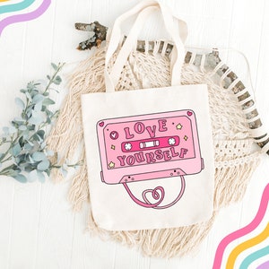 May include: A pink tote bag with a pink cassette tape design that says "LOVE YOURSELF" in white letters. The bag has white straps and is on a white wooden surface.