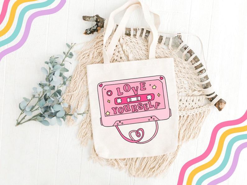 May include: A pink tote bag with a pink cassette tape design that says "LOVE YOURSELF" in white letters. The bag has white straps and is on a white wooden surface.