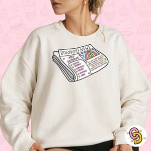 May include: A white sweatshirt featuring a graphic of a newspaper headline that reads "Breaking News You Deserve" with a rainbow and the words "Healing, Respect, Happiness, Care, Kindness, Love".