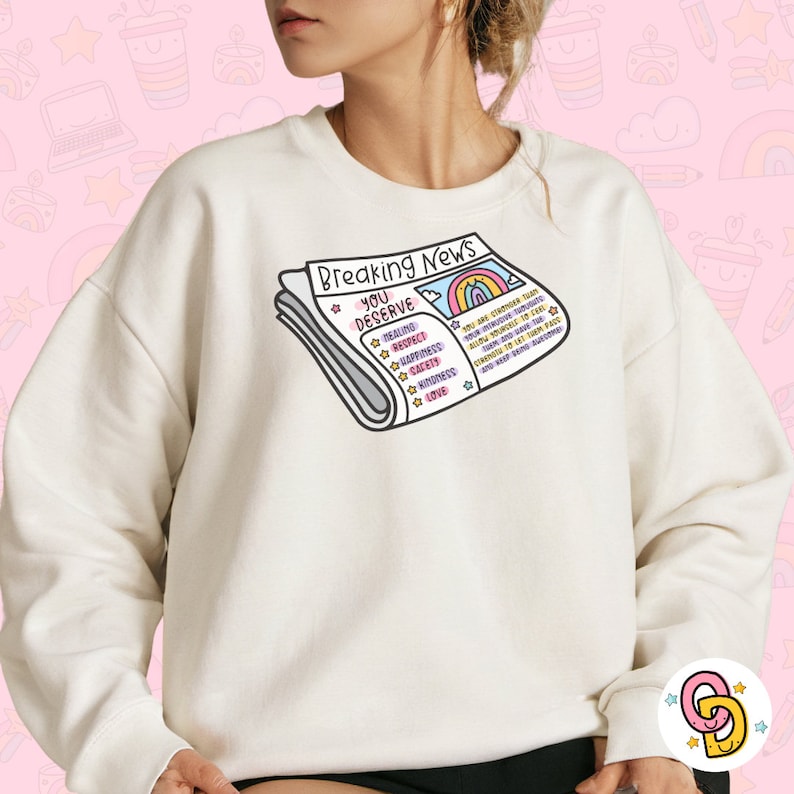 May include: A white sweatshirt featuring a graphic of a newspaper headline that reads "Breaking News You Deserve" with a rainbow and the words "Healing, Respect, Happiness, Care, Kindness, Love".