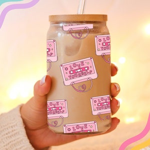 May include: A clear glass jar with a wooden lid and a straw. The jar is filled with a brown liquid and has a pink and white pattern of cassette tapes with the text "LOVE YOURSELF" on them.