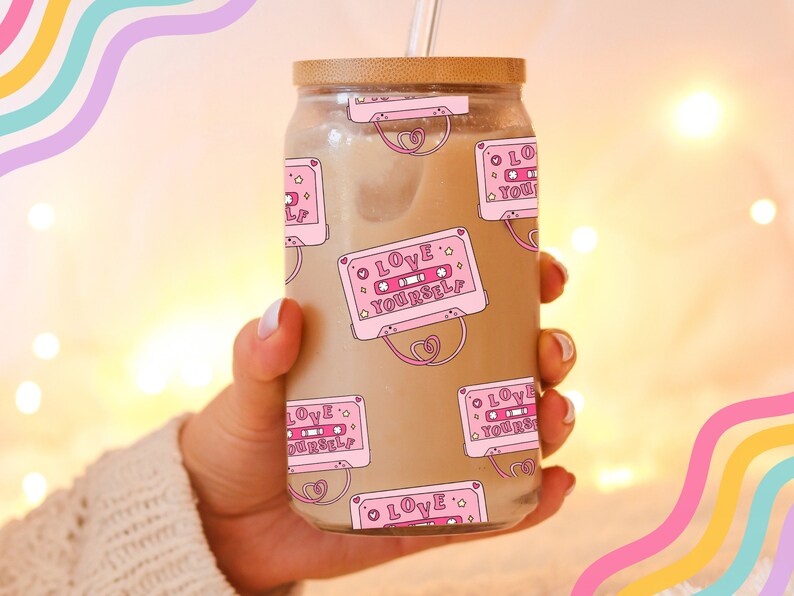 May include: A clear glass jar with a wooden lid and a straw. The jar is filled with a brown liquid and has a pink and white pattern of cassette tapes with the text "LOVE YOURSELF" on them.
