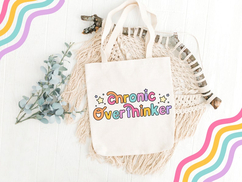 May include: A white tote bag with the text "Chronic Overthinker" in rainbow colors and yellow stars. The bag is on a white wooden surface with a branch and a macrame wall hanging.