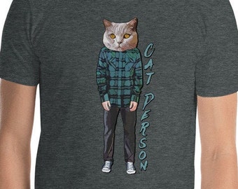 May include: A dark heather grey t-shirt featuring a cartoon cat wearing a plaid shirt and jeans. The text "CAT PERSON" is printed vertically on the right side of the shirt.