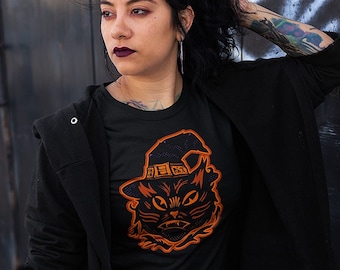 May include: Black t-shirt with an orange graphic of a black cat wearing a witch's hat. The cat has a spooky expression and the hat has stars on it. The text "Kryptic Society" is on the bottom of the image.