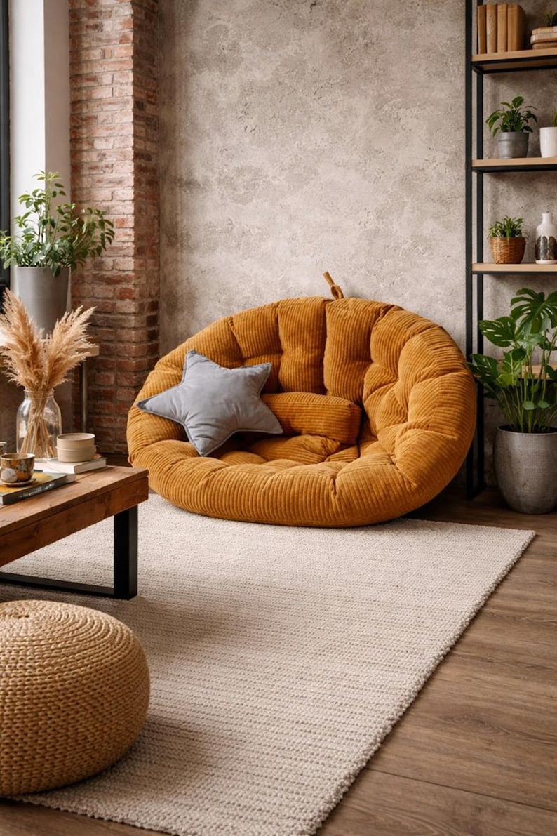 May include: A large, round, mustard-colored corduroy floor cushion with a gray star-shaped pillow and a smaller bolster pillow. The cushion is in a living room setting with a neutral rug and wooden furniture.