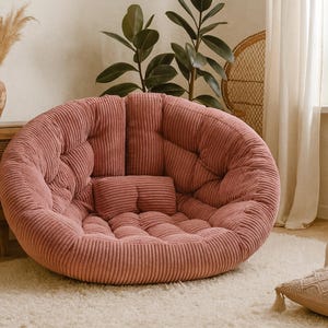 May include: A large, round, dusty rose corduroy floor chair with a matching small pillow. The chair is on a cream-colored rug. A wooden cabinet, framed artwork, and plants are in the background, creating a cozy, inviting atmosphere.