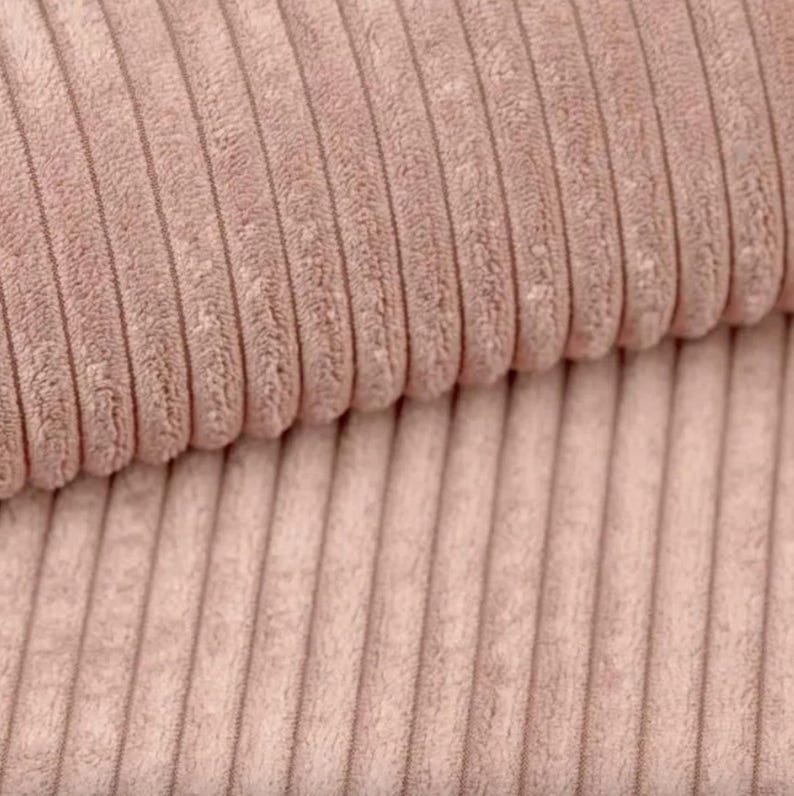May include: Close-up of a section of corduroy fabric in a soft, muted rose color. The fabric has a ribbed texture with vertical lines, creating a plush and tactile appearance. The corduroy is folded over, showing the texture and depth of the material.