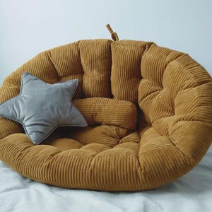 May include: A mustard-yellow corduroy floor cushion with a matching bolster pillow and a gray star-shaped pillow. The round cushion has a tufted design and a small loop at the top. The pillows add a decorative touch to the cozy seating.