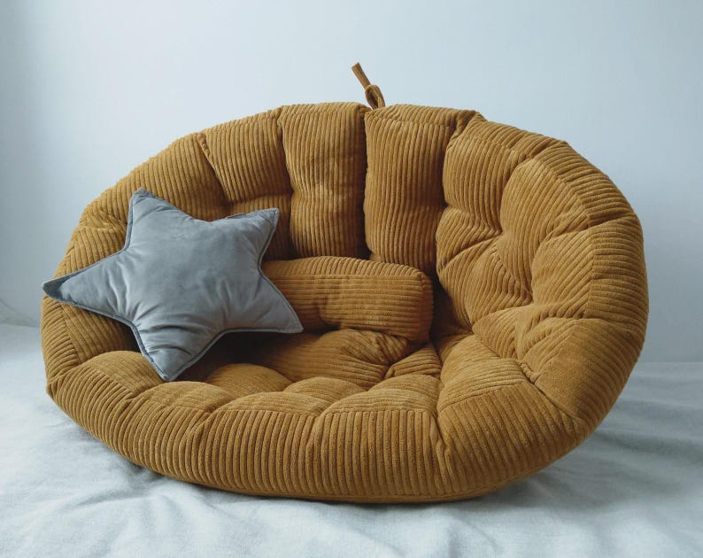 May include: A mustard-yellow corduroy floor cushion with a matching bolster pillow and a gray star-shaped pillow. The round cushion has a tufted design and a small loop at the top. The pillows add a decorative touch to the cozy seating.